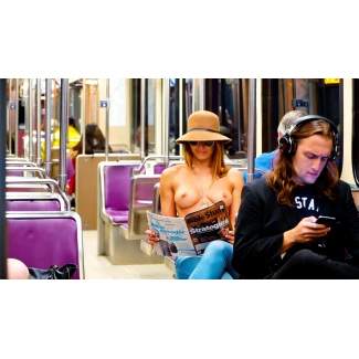 Topless woman wearing jeans a wide brimmed hat and sunglasses reads a magazine while showing her medium round breasts on a subway train