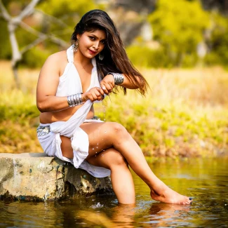 Shrutika Londhe seated on concrete slab in shallow water wearing white halter top and sheer skirt against blurred dry grass background