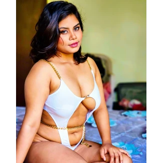 Desi woman with full breasts in white swimsuit with gold chain detailing seated on bed