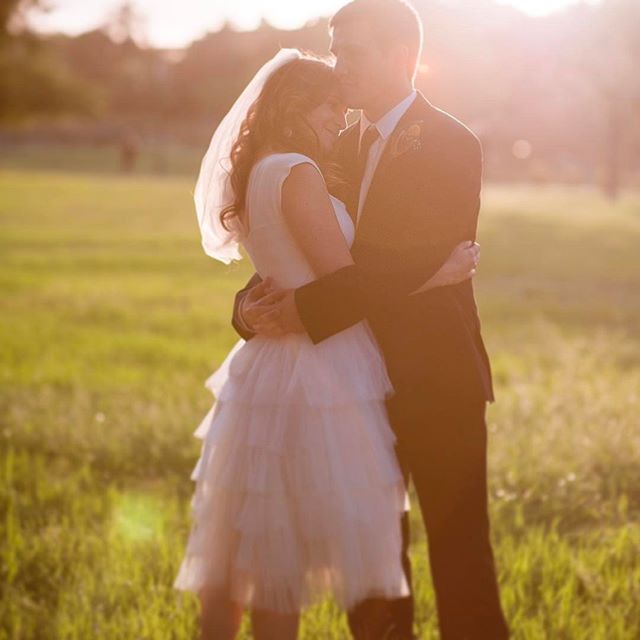 A bride and groom embrace in a field during their wedding, backlit by a warm sunset.
