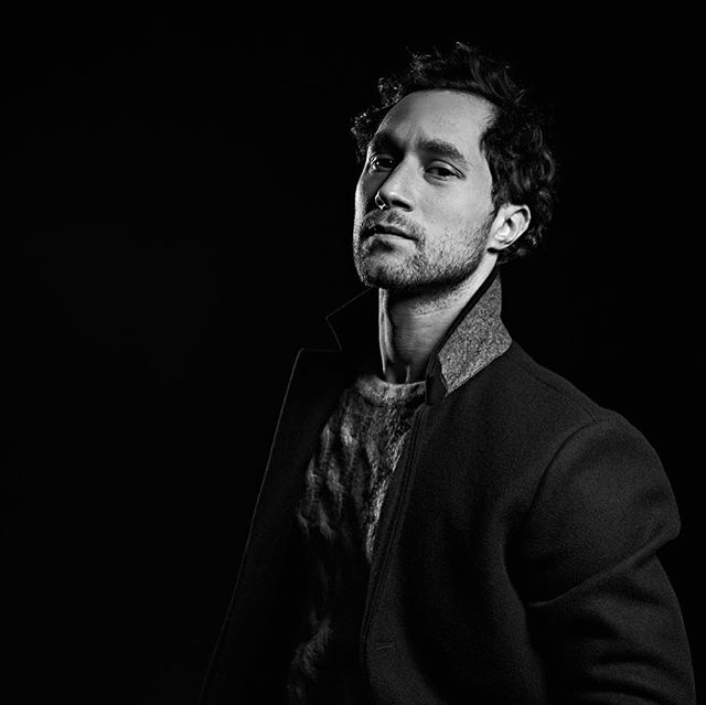 A stylish man poses in a black and white studio portrait with dramatic lighting, showcasing a confident and serious demeanor.