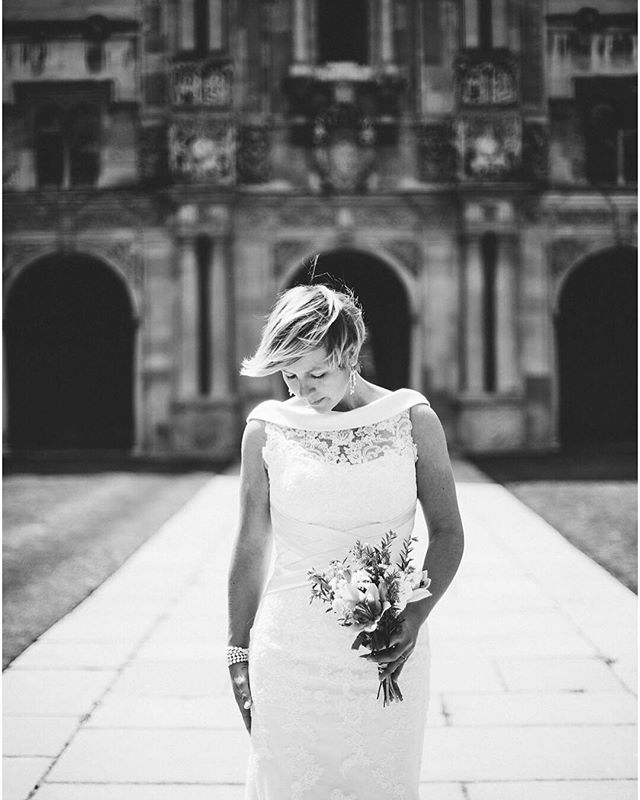 A bride in a lace wedding dress holds a bouquet in front of an old building in this black and white photo.