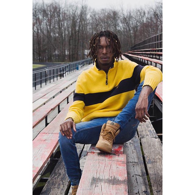 A stylish man with dreadlocks sitting on bleachers in a yellow sweater, jeans, and boots.