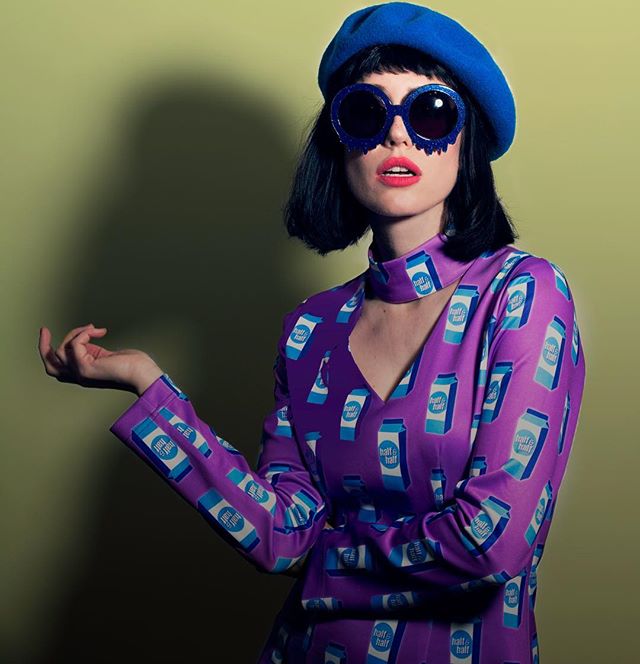 A fashionable woman poses in a purple half-and-half carton print dress, blue beret, and statement sunglasses.