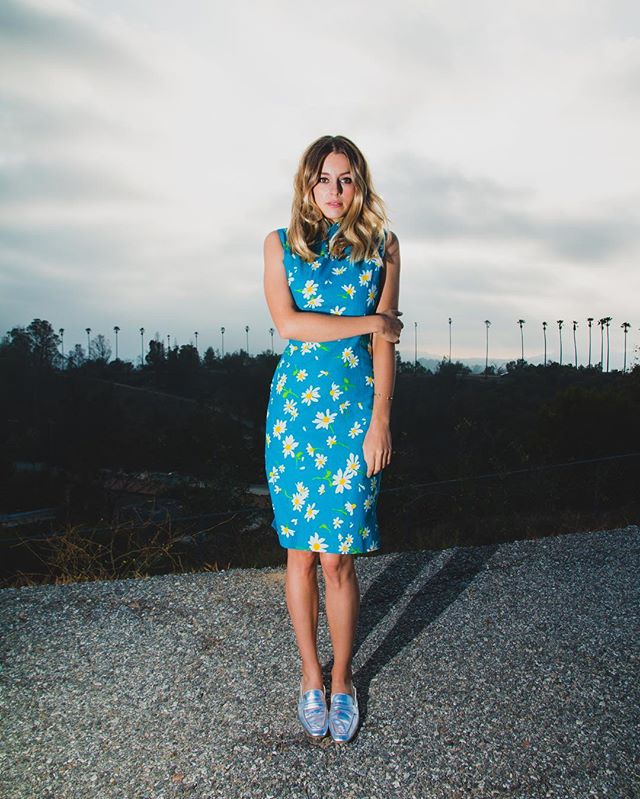 A woman in a blue floral dress stands on a road. She has her arms crossed and is looking at the camera.