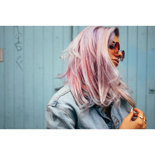 A stylish woman with pastel pink hair and sunglasses poses outdoors against a blue wall in a denim jacket.