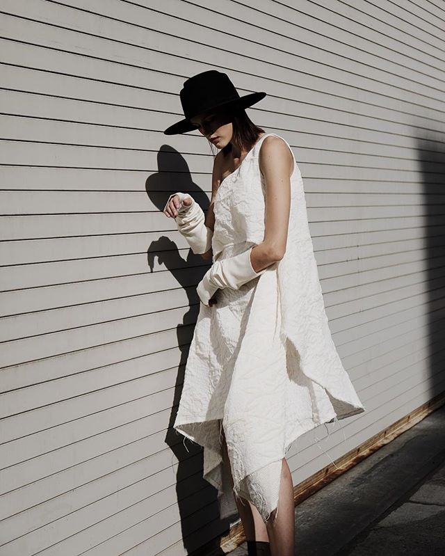 A woman in a white dress and black hat poses against a gray wall in an editorial fashion setting.