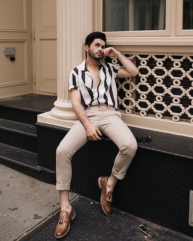 A stylish man in a striped shirt and khaki pants sits thoughtfully in an urban setting.