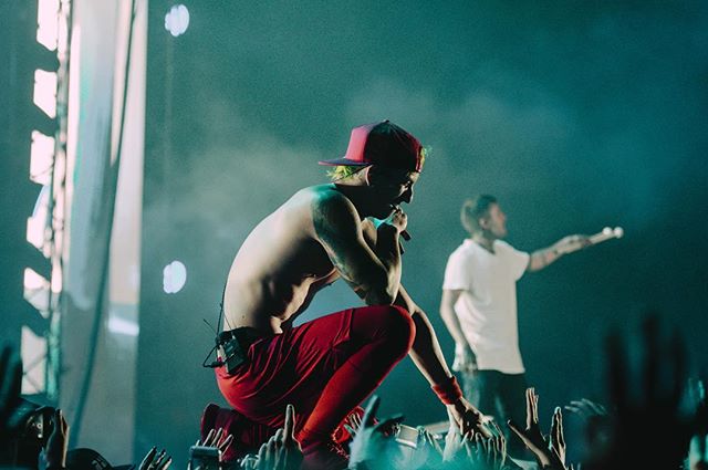 A shirtless singer in a red hat kneels on stage during a concert, performing for a large crowd.