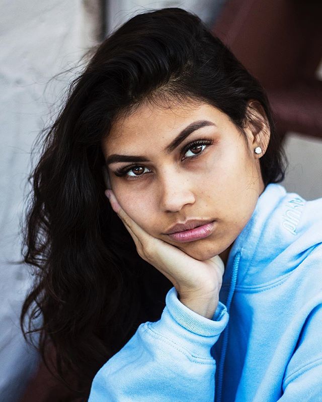 A young woman with brown hair poses thoughtfully with her hand on her face, wearing a blue hoodie.