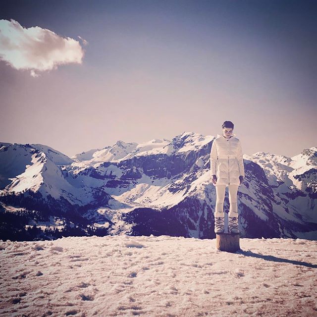A woman in winter apparel stands on a snowy mountain top with snow-capped mountains in the background.