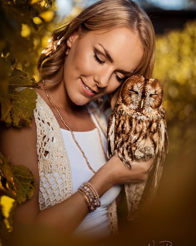 A blonde woman gently holds a brown owl with closed eyes in a peaceful, sun-drenched outdoor setting.