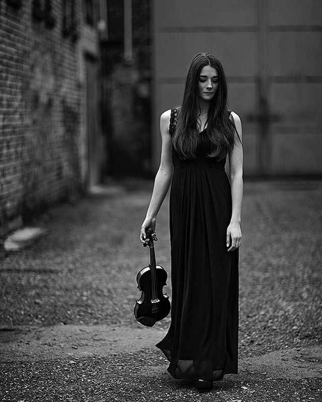 A woman in a black dress walks in an alleyway holding a violin, creating an emotional and artistic image.