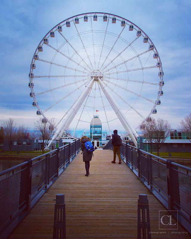 A Ferris wheel towers over people strolling on a bridge in a scenic urban landscape.