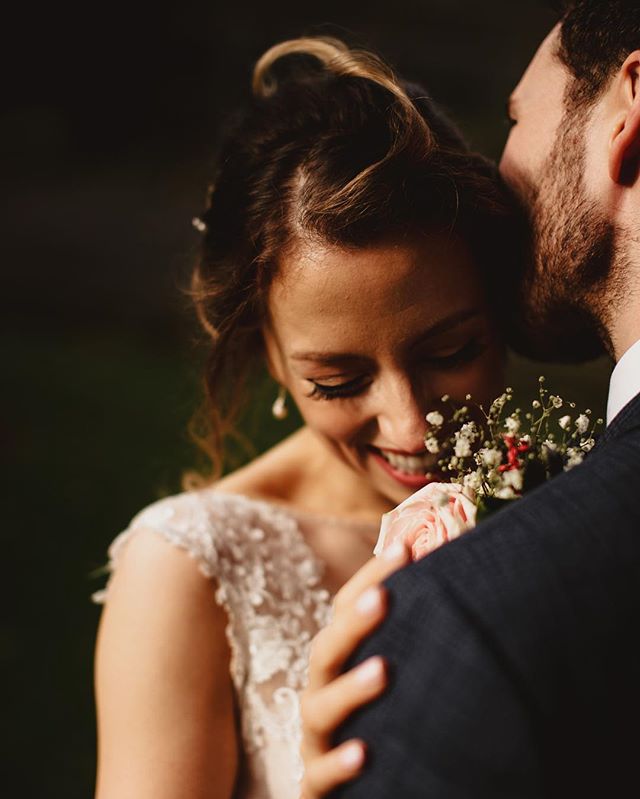 A bride smiles tenderly as the groom embraces her, holding a beautiful flower bouquet in an intimate wedding moment.