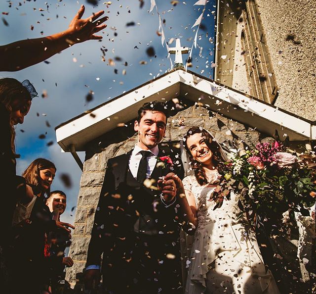 A happy bride and groom exit the church under a shower of confetti, surrounded by wedding guests.