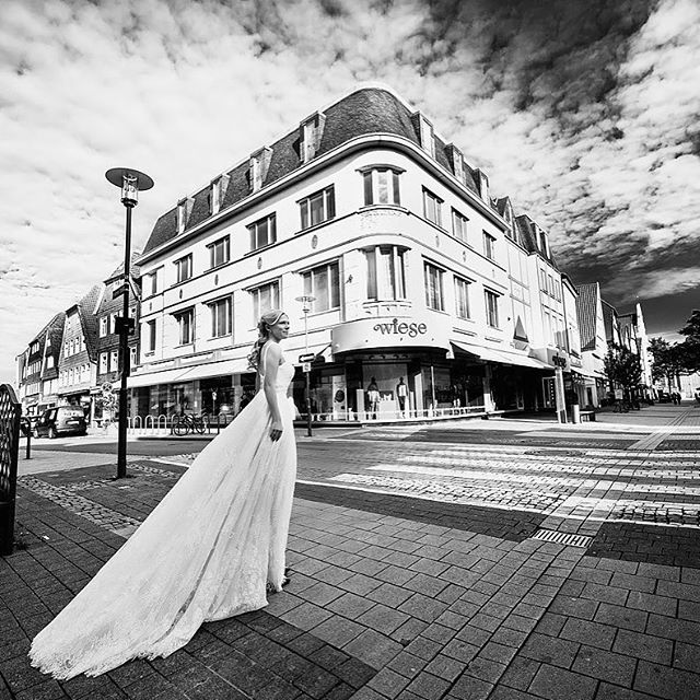 A bride in a white dress walks on a city street in a black and white photograph.