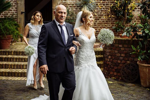 A bride is escorted by her father and bridesmaid at an outdoor wedding, creating a heartwarming and elegant moment.