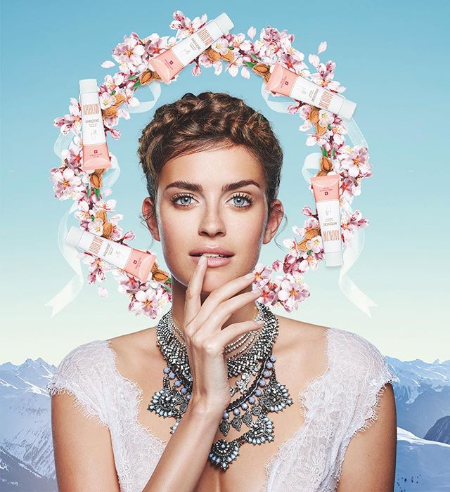 A beautiful woman wears a flower crown and necklace while promoting beauty products in a serene, elegant outdoor setting.