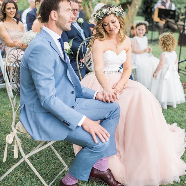 A bride and groom hold hands during their outdoor wedding ceremony, surrounded by guests and flower girls.