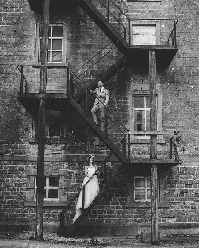 A bride and groom stand on an outdoor fire escape staircase in a black and white wedding photo.
