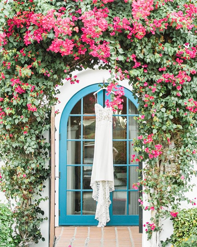 A wedding dress hangs in a doorway framed by vibrant pink bougainvillea, creating a romantic and elegant scene.