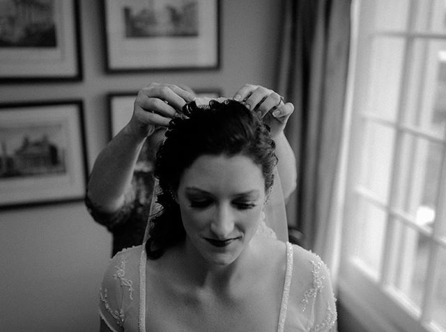 A bride getting ready for her wedding, having her veil adjusted indoors near a window in a black and white photo.