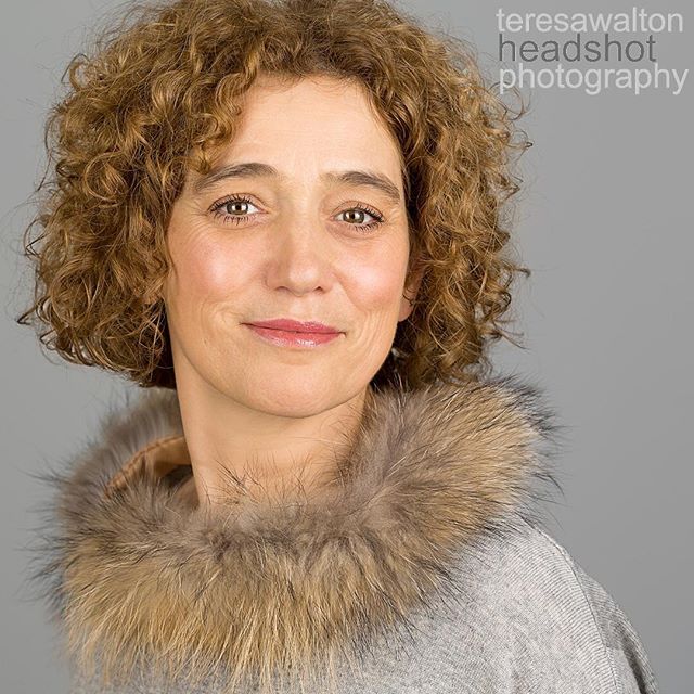 Professional headshot of a woman with curly hair in a fur-lined coat, smiling softly against a gray background.