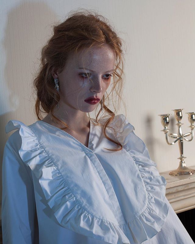 A redhead woman poses in a ruffled white dress and earrings for an elegant editorial fashion shoot.