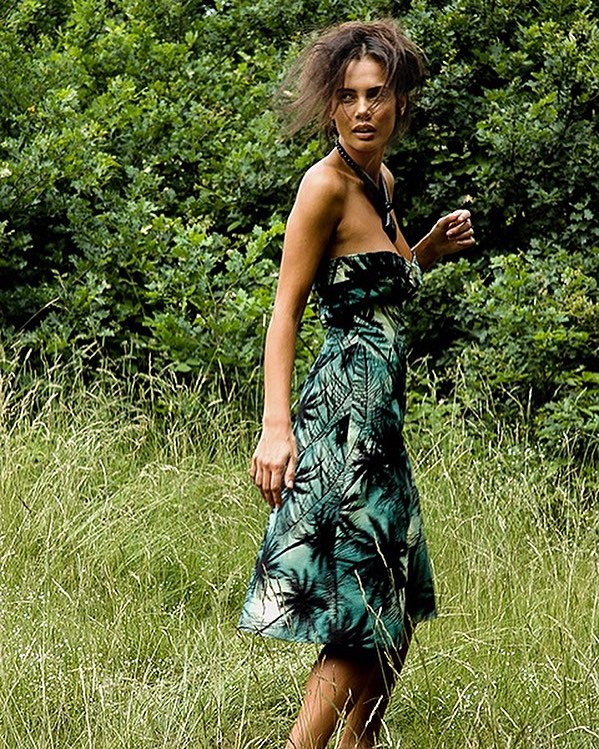 A woman in a tropical print dress poses in a grassy field with a forest backdrop.