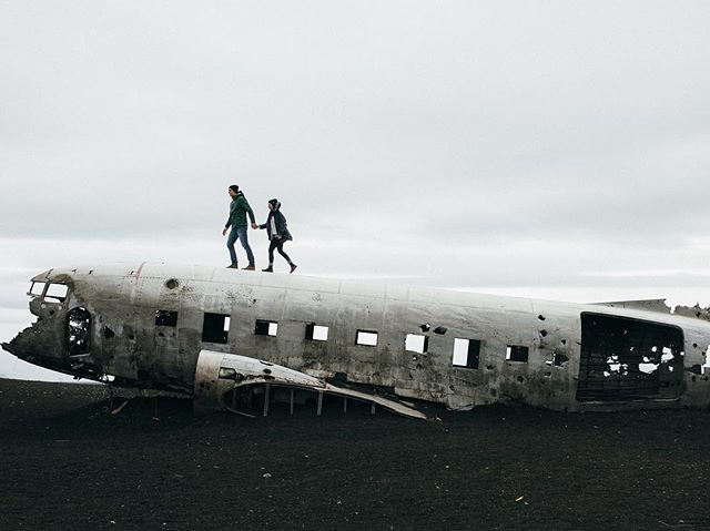 A man and woman walk along the wreckage of a plane on a black sand beach in Iceland.