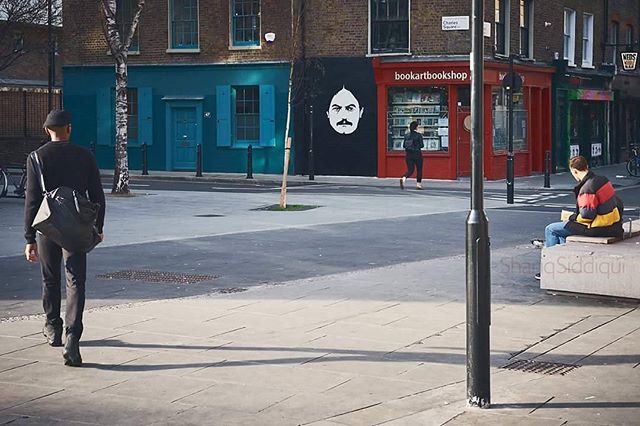 A man walks on a city street with a bookstore and mural in the background on a sunny day.