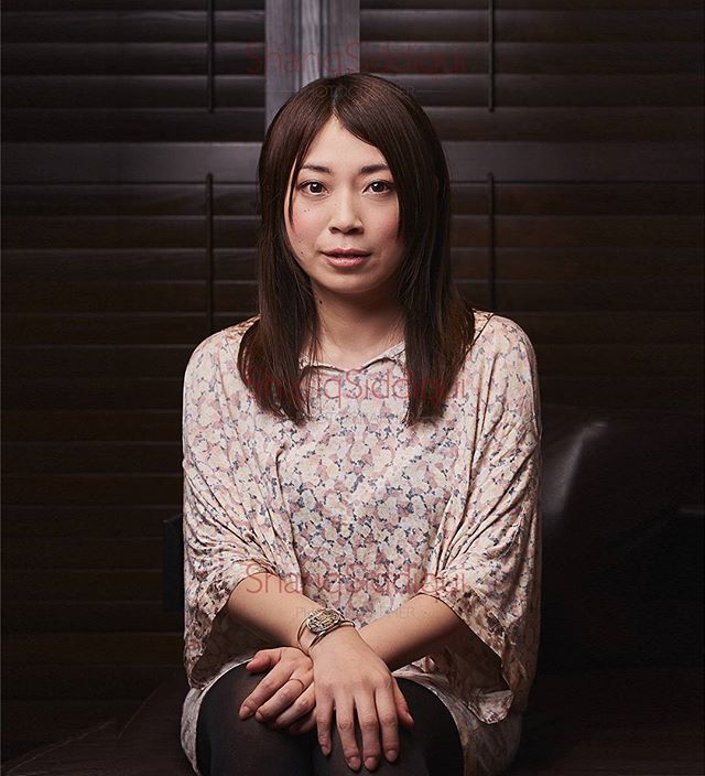Portrait of an Asian woman with a floral top sitting indoors against a dark background.