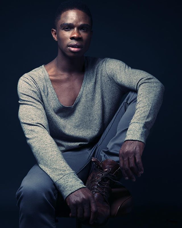 A man sits in a studio wearing a gray shirt and brown boots in a modern editorial style.