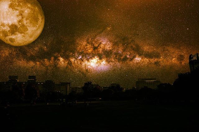 A surreal cityscape is illuminated by a golden moon and the Milky Way on a starry night.