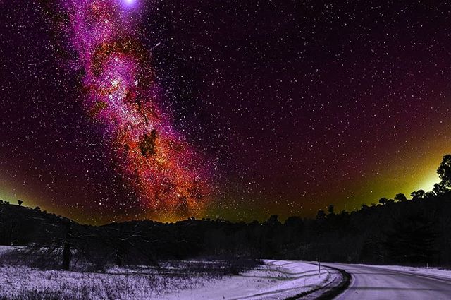 A vibrant, long exposure image shows the night sky filled with stars above a snowy landscape with a winding road.