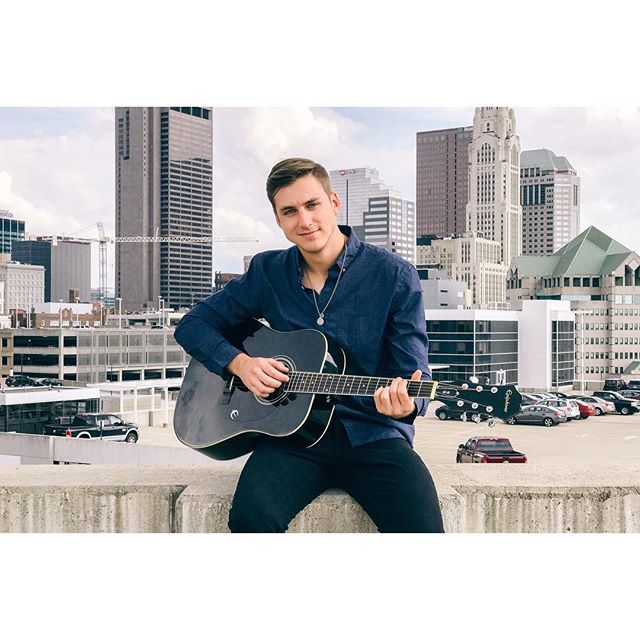 A young man sits on a rooftop playing guitar against a city skyline.