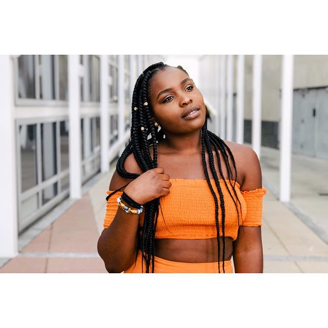 A beautiful young woman in an orange top and braids looks confidently into the distance.