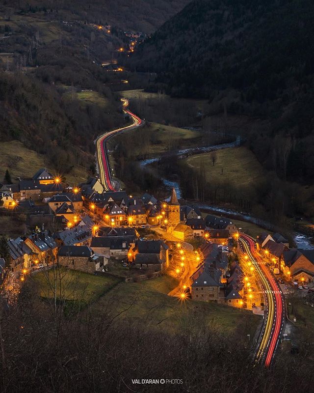 Night view of a small village nestled in a mountain valley with light trails on the road.