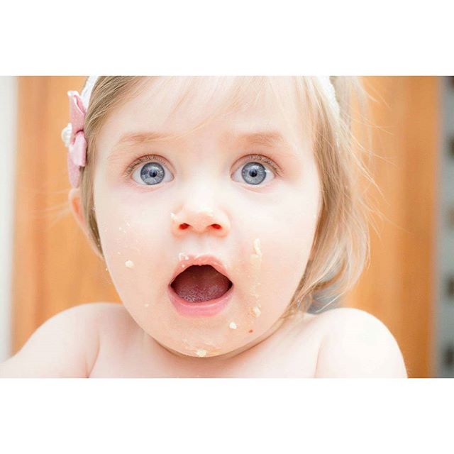 A cute baby girl with an open mouth and food smeared on her face looks surprised and happy.