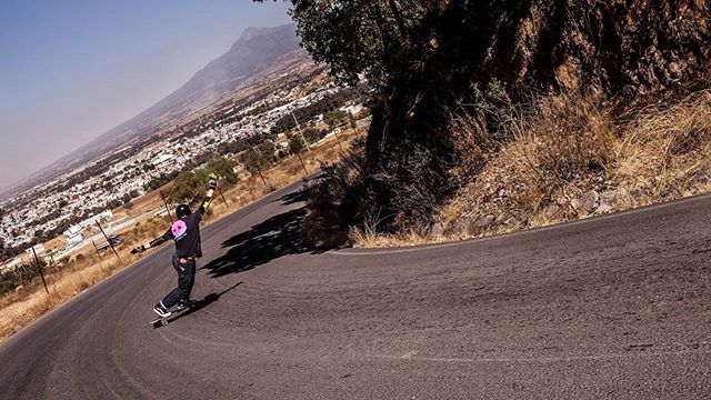 A man skateboards downhill on a winding road with outstretched arms and a city view.