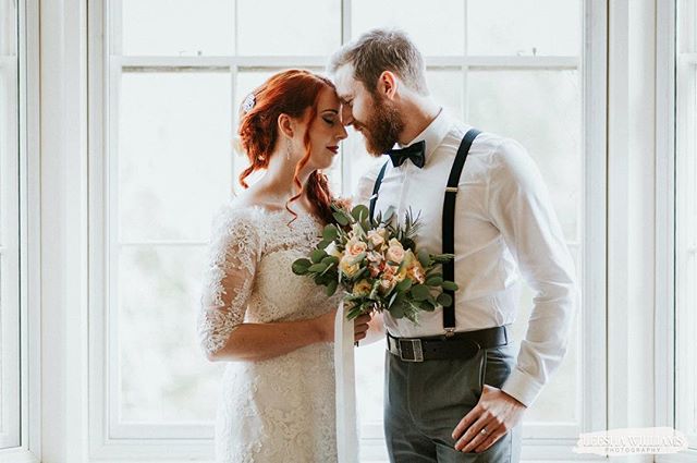 A bride and groom share an intimate moment in front of a window, holding a bouquet during their wedding day.