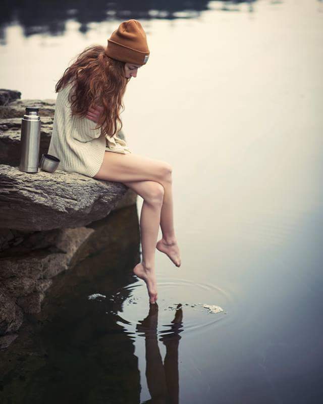 A serene woman in a beanie touches the water while sitting on a rock near a lake with a thermos.