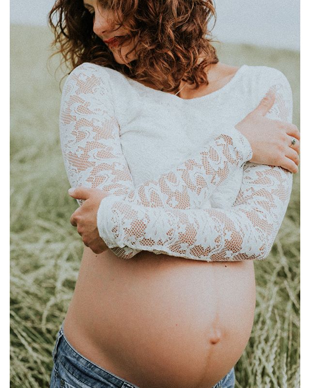 A pregnant woman gently cradles her baby bump in a serene outdoor setting, wearing a white lace top and jeans.