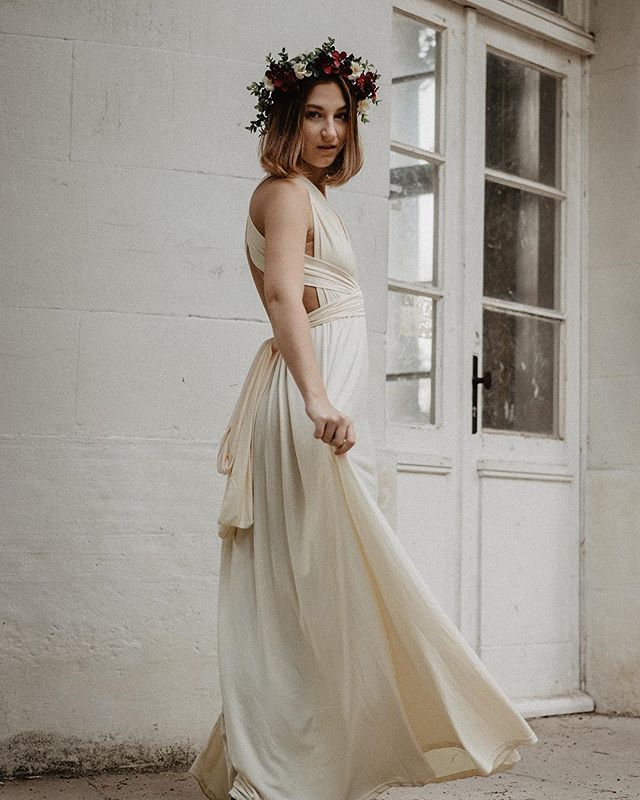 A woman in a flowing gown and flower crown poses serenely by a doorway in soft, dreamy light.