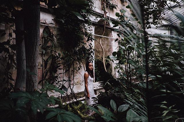 A woman in a white dress stands among lush plants in a vintage greenhouse.