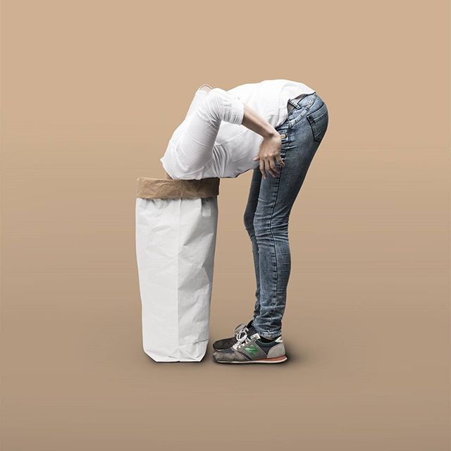 A person with their head in a paper trash can against a beige backdrop in a conceptual studio shot.