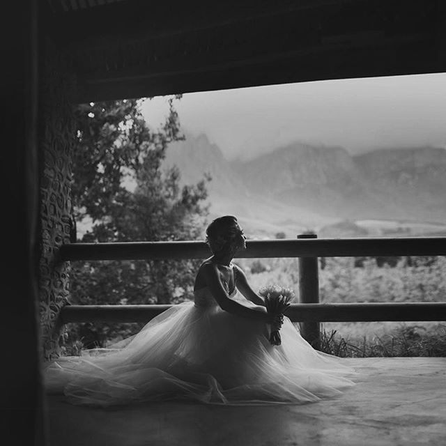 A bride sits in her dress on a balcony overlooking mountains, holding her bouquet.