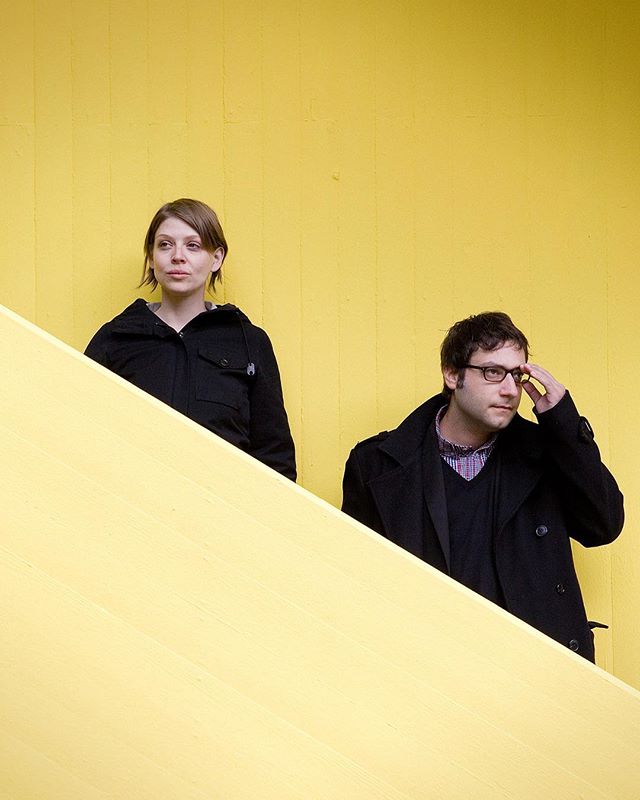 A serious looking couple poses against a bright yellow wall in a modern, editorial style.