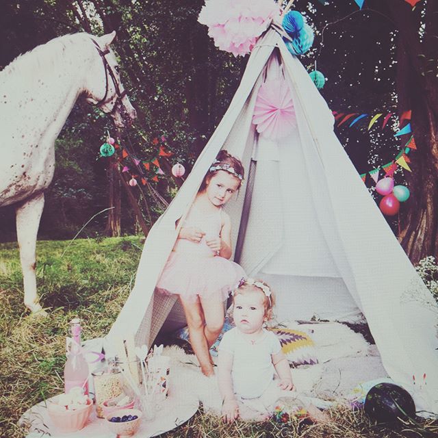 Two young girls in tutus are in a teepee with a white horse nearby, celebrating outdoors with balloons and decorations.
